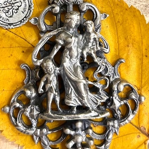 May include: A silver pendant featuring a woman and two children, with ornate scrollwork and a crown-like top. The pendant hangs from a black cord and has a tag that reads "Made in Germany". The pendant is set against a yellow leaf.