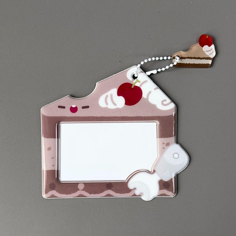 May include: A novelty ID badge holder shaped like a slice of cake, featuring a cherry on top and a bite taken out. The holder is light brown with white frosting accents and a white rectangular window for an ID card. A small cake slice charm hangs from a chain.