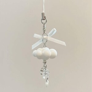 May include: A white cloud charm with a small white bow and a silver-coloured clasp. The charm has a string for hanging and features a cluster of white orbs and a dangling crystal accent.