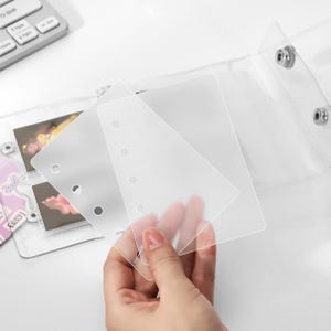 Può includere: Raccoglitore in plastica trasparente con chiusura a scatto, contenente diverse piccole foto. Il raccoglitore ha alcuni inserti in plastica trasparente smerigliata. Il raccoglitore è su una superficie bianca.