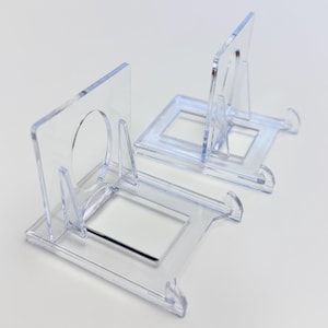 May include: Two clear acrylic display stands. Each stand has a rectangular base with a square opening and a clear, upright support with a circular opening. The stands are designed to hold small items.