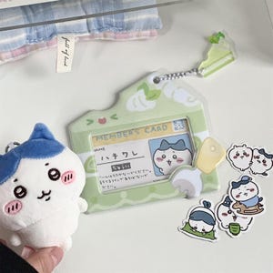 May include: A collection of cute, cartoon-style accessories. Includes a light green member's card holder with a cartoon character, a plush toy with a blue and white design, and several stickers. The card holder has Japanese text.