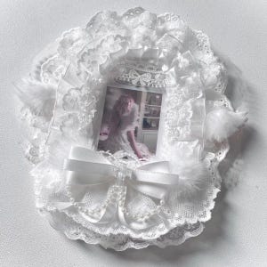 May include: A white circular photo frame decorated with layers of ruffled lace, sheer fabric, and soft faux fur accents. A satin ribbon bow sits at the bottom, adorned with a delicate string of white faux pearls surrounding a central photograph.