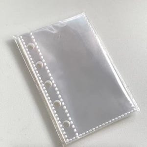 Puede incluir: Una funda transparente de plástico para encuadernación con múltiples agujeros a lo largo del borde izquierdo para su inserción en una carpeta. La funda es rectangular y está sellada por tres lados, con una pequeña abertura en la parte superior. Diseñada para guardar objetos pequeños.