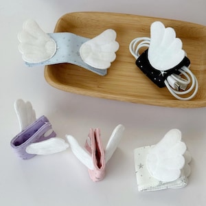 May include: A collection of angel wing-shaped cable organizers in various colors, including blue, black, purple, pink, and white. Each organizer features white felt wings and is designed to hold and manage cords. Some have star patterns.