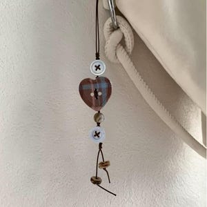May include: A decorative charm featuring a heart-shaped button with a plaid design in shades of brown and blue. The charm also includes white buttons with black cross details, and small, round beads. The charm is attached to a dark brown cord.