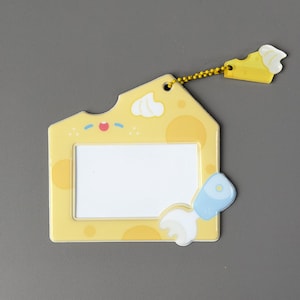 May include: A yellow cheese-shaped ID badge holder with a white rectangular window. The holder has a small cheese slice charm attached by a gold chain. Decorative elements include a smiling face and a blue and white brush.