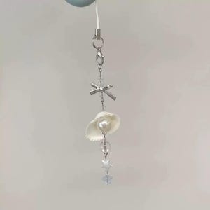 May include: A delicate, silver-toned charm featuring a miniature white seashell hat with a pearl, clear beads, and star-shaped accents. The charm is suspended from a silver chain and clasp, perfect for a necklace or keychain.
