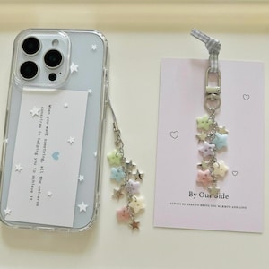 Puede incluir: Funda de teléfono transparente con un fondo azul claro y estrellas blancas. Un charm plateado con cuentas en forma de estrella pastel y estrellas de metal cuelga de la funda. Una tarjeta con el texto "By Our Side" y corazones también está en la imagen.
