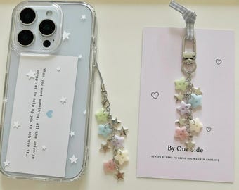 May include: A clear phone case with a light blue backing and white star details. A silver charm with pastel star-shaped beads and metal stars hangs from the case. A card with the text "By Our Side" and hearts is also visible.