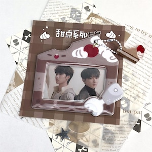 May include: A brown photo frame shaped like a house with a cherry and cake charm. The frame holds two photos of young men. The text "甜点系列安" is visible on the frame. The background includes printed paper.