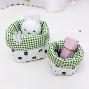 Puede incluir: Dos pequeñas cestas de tela con estampados de cuadros verdes y tréboles. Una cesta contiene un peluche de conejo blanco y la otra un labial rosa. Las cestas están sobre una superficie blanca, con una pegatina de trébol verde y el texto "#clover".