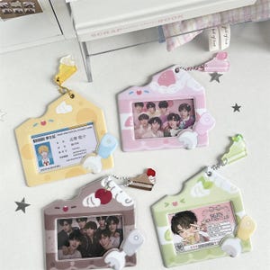 May include: Four decorative ID badge holders in pastel colors. Each holder has a house-shaped design with a photo or ID card inside. The holders are yellow, pink, brown, and green, with decorative charms and keychains.