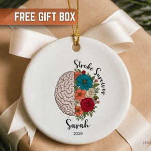 May include: White ceramic ornament with a gold string, featuring a brain illustration and colorful flowers. Text reads "Stroke Survivor," the name "Sarah," and the year "2026."