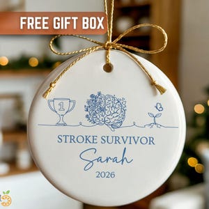 May include: A white ceramic ornament with a gold string for hanging. The ornament features a blue line drawing of a trophy, brain, and plant. Text on the ornament reads "STROKE SURVIVOR" and "Sarah 2026". A gold bow is at the top.