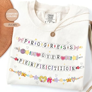 May include: Ivory-colored t-shirt with the words "PROGRESS OVER PERFECTION" in a playful, colorful design. The design includes hearts, stars, butterflies, and pencils. A gold necklace is draped over the shirt.