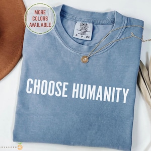 May include: A light blue t-shirt with the words "CHOOSE HUMANITY" in white block letters. The shirt has a crew neck and short sleeves. A gold necklace with a pendant is draped on the shirt. The shirt is labeled "Comfort Colors".
