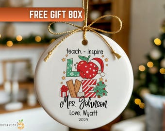 May include: White ceramic Christmas ornament with a gold string. The ornament features a colorful design with an apple wearing a Santa hat, the word "LOVE", a Christmas tree, and the text "teach - inspire". The ornament is personalized with a name and the year 2025.