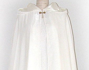 Puede incluir: Una capa de color marfil con capucha, exhibida en un maniquí. La capa, de largo hasta el suelo, está hecha de una tela fluida y tiene un cierre decorativo en el cuello. El diseño sugiere una prenda formal o de novia.