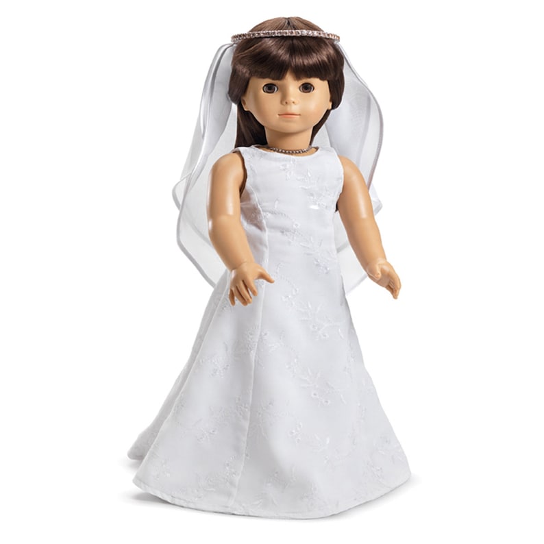 May include: A white doll wearing a white bridal gown with a veil and a crown. The doll has brown hair and blue eyes.