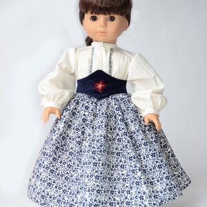 May include: A doll wearing a white blouse with a blue and white floral skirt and a blue sash with a red flower accent.