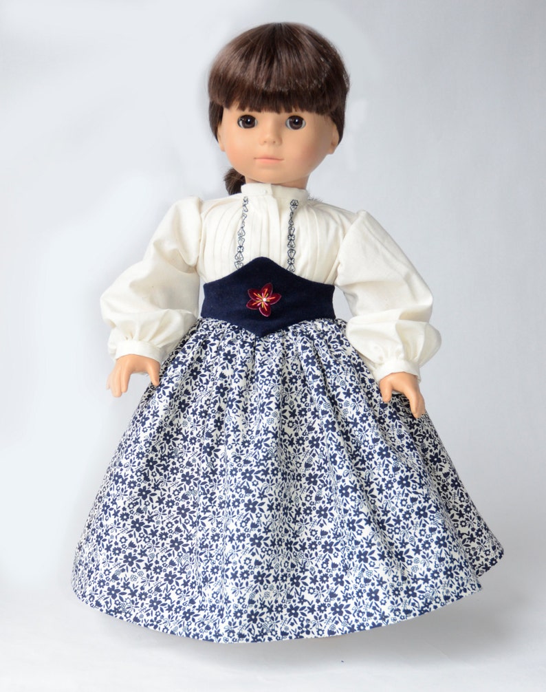 May include: A doll wearing a white blouse with a blue and white floral skirt and a blue sash with a red flower accent.