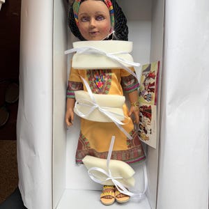 Puede incluir: Mu&ntilde;eca en una caja, con un vestido amarillo con bordados coloridos y sandalias a juego. La mu&ntilde;eca tiene un pa&ntilde;uelo en la cabeza y un velo negro. Est&aacute; asegurada con espuma y cinta blancas.
