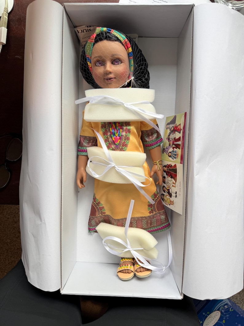 Puede incluir: Mu&ntilde;eca en una caja, con un vestido amarillo con bordados coloridos y sandalias a juego. La mu&ntilde;eca tiene un pa&ntilde;uelo en la cabeza y un velo negro. Est&aacute; asegurada con espuma y cinta blancas.