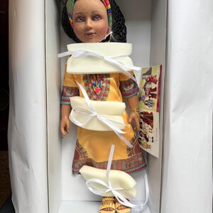 Puede incluir: Mu&ntilde;eca en una caja blanca, con un vestido amarillo con patrones geom&eacute;tricos coloridos. La mu&ntilde;eca tiene un velo de red negro, una diadema colorida y sandalias amarillas. La mu&ntilde;eca est&aacute; asegurada con relleno de espuma blanca y cintas blancas.