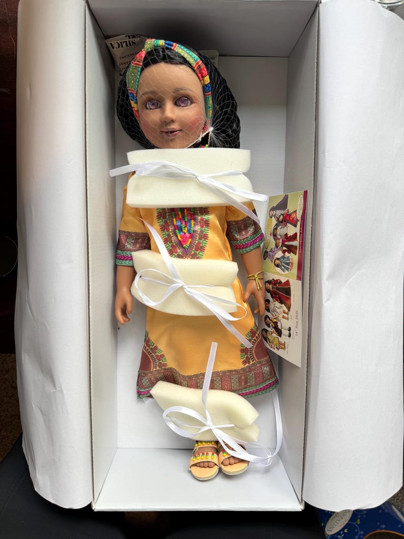 Puede incluir: Mu&ntilde;eca en una caja blanca, con un vestido amarillo con patrones geom&eacute;tricos coloridos. La mu&ntilde;eca tiene un velo de red negro, una diadema colorida y sandalias amarillas. La mu&ntilde;eca est&aacute; asegurada con relleno de espuma blanca y cintas blancas.