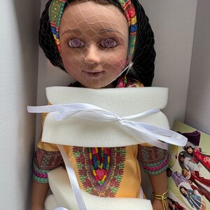 Puede incluir: Mu&ntilde;eca con cabello oscuro, vestida con un vestido amarillo con un dise&ntilde;o bordado colorido. La mu&ntilde;eca tiene una diadema estampada y un velo. La mu&ntilde;eca est&aacute; empaquetada en una caja con espuma protectora y cintas blancas.
