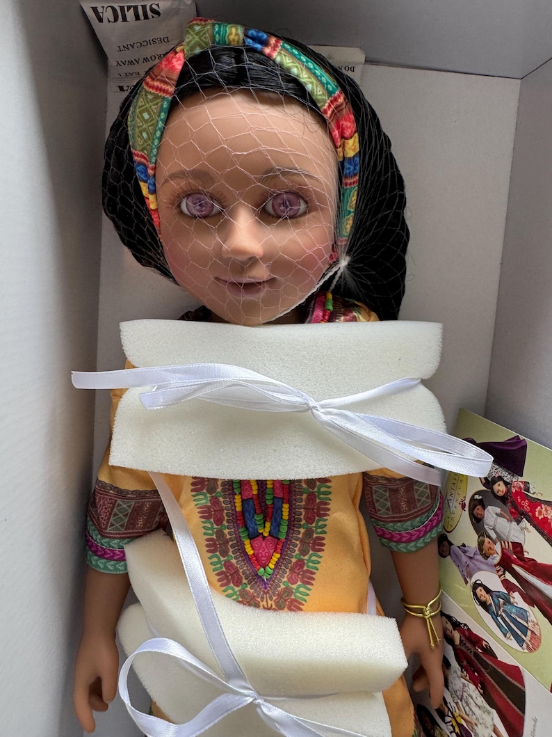 Puede incluir: Mu&ntilde;eca con cabello oscuro, vestida con un vestido amarillo con un dise&ntilde;o bordado colorido. La mu&ntilde;eca tiene una diadema estampada y un velo. La mu&ntilde;eca est&aacute; empaquetada en una caja con espuma protectora y cintas blancas.