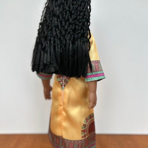 Puede incluir: Una mu&ntilde;eca con largo cabello negro trenzado, vestida con un vestido amarillo dorado con ribete estampado colorido. El vestido tiene cuello redondo y mangas largas. La mu&ntilde;eca est&aacute; de espaldas.