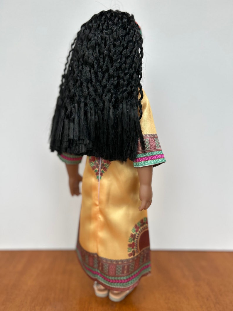 Puede incluir: Una mu&ntilde;eca con largo cabello negro trenzado, vestida con un vestido amarillo dorado con ribete estampado colorido. El vestido tiene cuello redondo y mangas largas. La mu&ntilde;eca est&aacute; de espaldas.