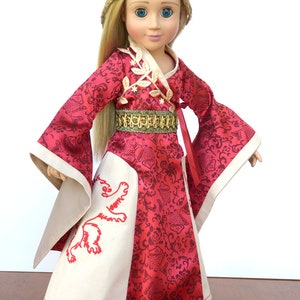 May include: A doll wearing a red and white dress with a gold belt and a lion embroidered on the skirt. The doll has long blonde hair and blue eyes.