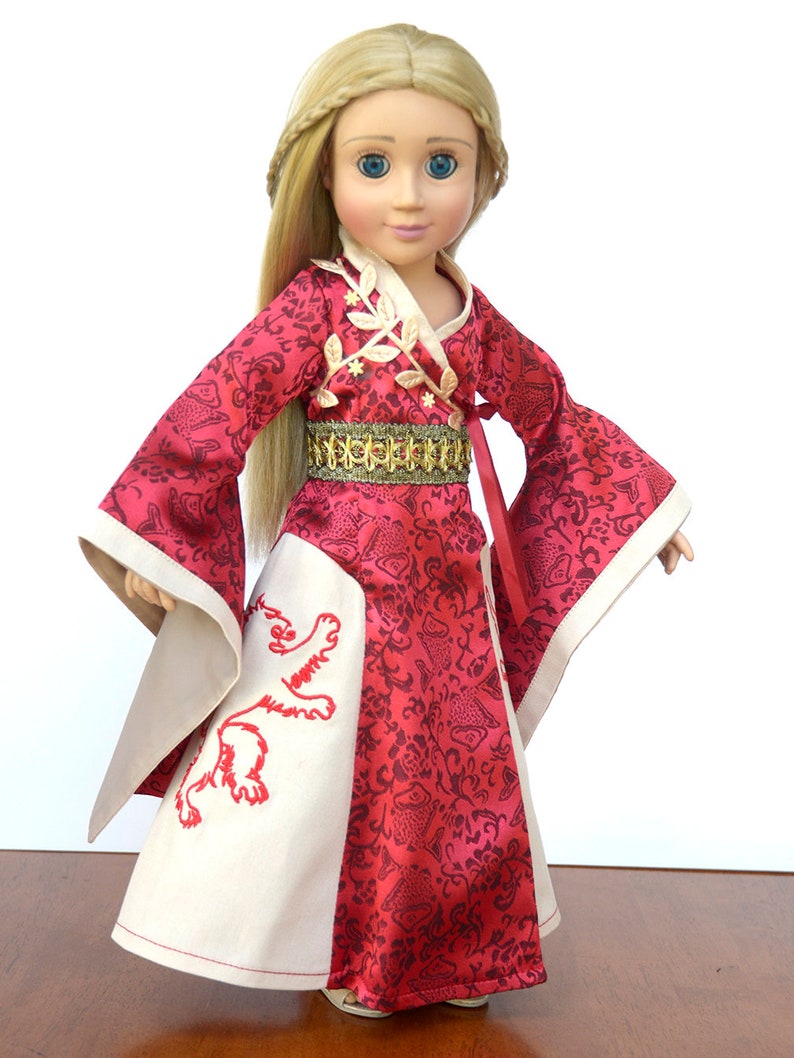May include: A doll wearing a red and white dress with a gold belt and a lion embroidered on the skirt. The doll has long blonde hair and blue eyes.