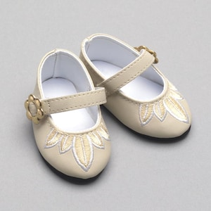 May include: A pair of cream-colored doll shoes with a floral embroidered design on the toe. The shoes have a strap with a gold-colored flower-shaped buckle. The soles are black, and the interior is white.