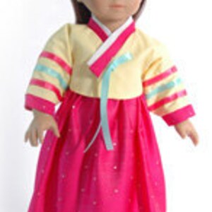 May include: A doll wearing a traditional Korean hanbok dress. The dress features a pale yellow top with pink trim and sleeves adorned with blue and pink ribbons. The long, bright pink skirt has a decorative gold border. The doll has brown hair and a small tiara.