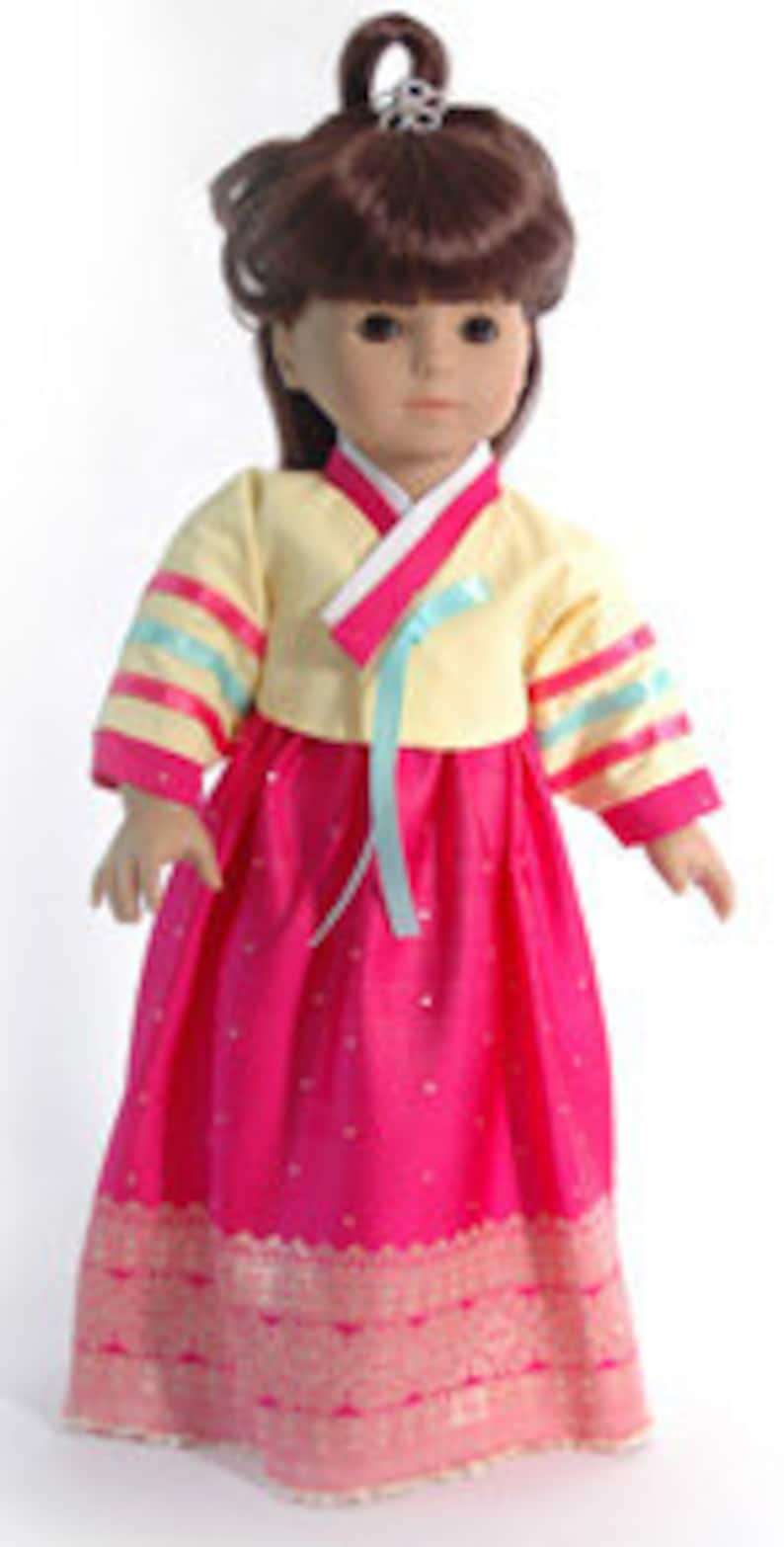 May include: A doll wearing a traditional Korean hanbok dress. The dress features a pale yellow top with pink trim and sleeves adorned with blue and pink ribbons. The long, bright pink skirt has a decorative gold border. The doll has brown hair and a small tiara.