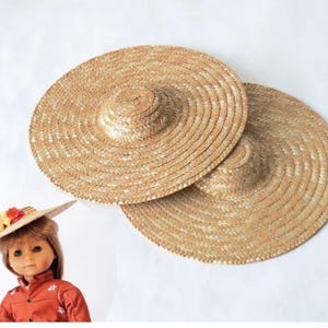 May include: Two round, flat-brimmed straw hats with a woven texture and a small, raised centre crown. A doll with brown hair wears a similar straw hat adorned with small red, yellow, and orange fabric flowers, paired with a rust-coloured shirt.