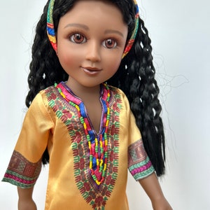 Puede incluir: Mu&ntilde;eca con piel oscura, ojos marrones y pelo largo y rizado negro. La mu&ntilde;eca lleva un vestido amarillo con adornos de cuentas de colores y una diadema estampada. El vestido tiene patrones decorativos en las mangas.