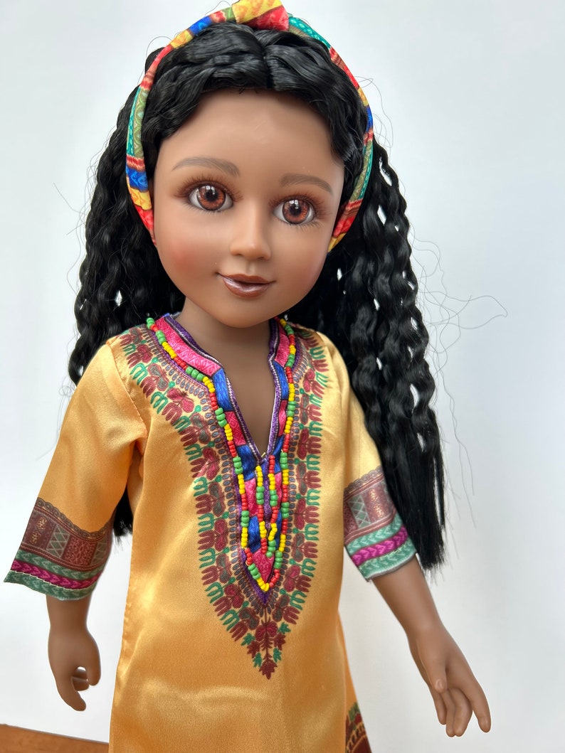 Puede incluir: Mu&ntilde;eca con piel oscura, ojos marrones y pelo largo y rizado negro. La mu&ntilde;eca lleva un vestido amarillo con adornos de cuentas de colores y una diadema estampada. El vestido tiene patrones decorativos en las mangas.