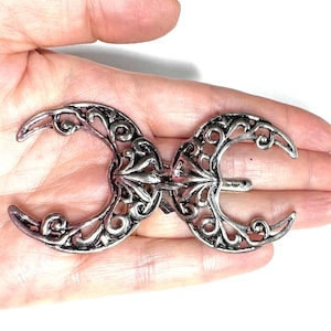 Può includere: Fermaglio per capelli in metallo argentato con un design simmetrico di due lune crescenti collegate al centro. Le lune presentano intricati motivi traforati e una barra decorativa per fissare il fermaglio.