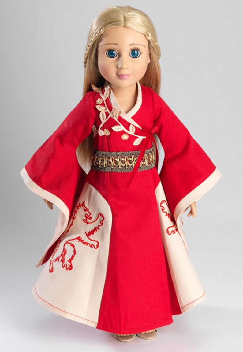 May include: A red and white doll dress with gold embroidery and a lion design. The doll is wearing a gold belt and has blonde hair and blue eyes.