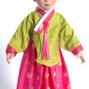 May include: A doll dressed in a traditional Korean hanbok. The doll's outfit includes a lime green top with long sleeves and a bright pink skirt. The skirt has a gold floral pattern. The doll has black hair and is wearing a white headpiece.