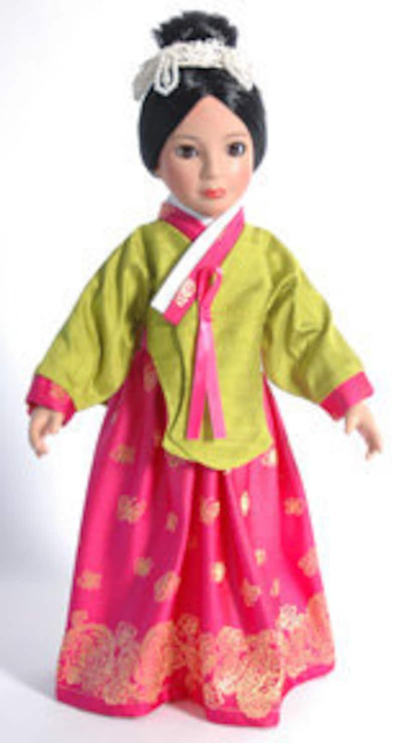 May include: A doll dressed in a traditional Korean hanbok. The doll's outfit includes a lime green top with long sleeves and a bright pink skirt. The skirt has a gold floral pattern. The doll has black hair and is wearing a white headpiece.