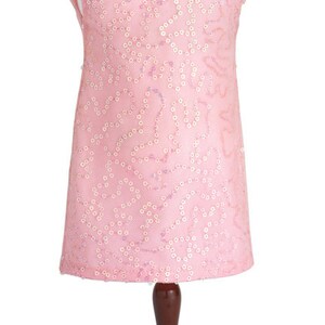 Pu&ograve; includere: Un abito rosa senza maniche con un motivo di paillettes e una spilla gioiello sul d&eacute;collet&eacute;. L'abito &egrave; esposto su un manichino di legno.