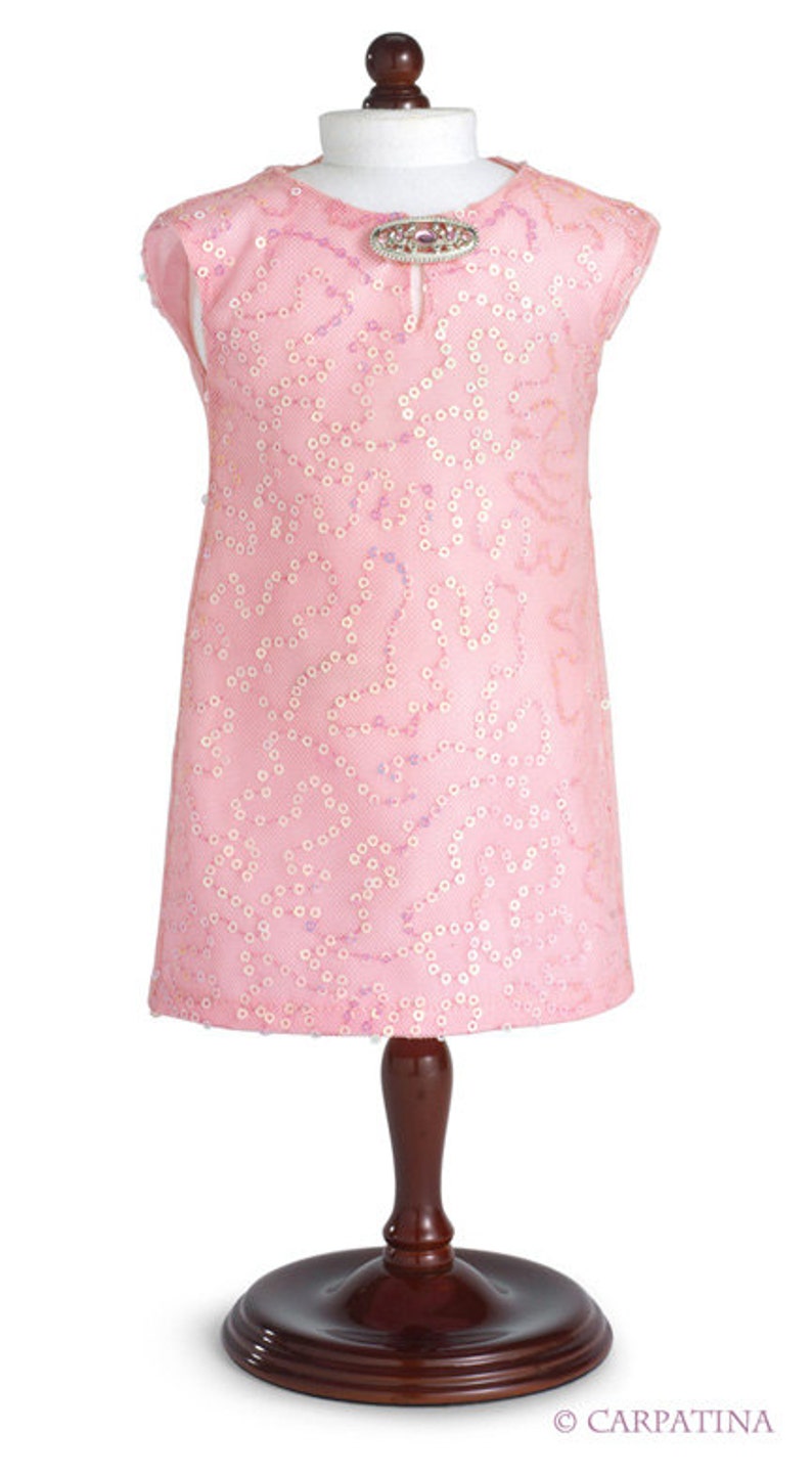 Pu&ograve; includere: Un abito rosa senza maniche con un motivo di paillettes e una spilla gioiello sul d&eacute;collet&eacute;. L'abito &egrave; esposto su un manichino di legno.