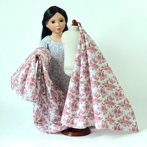 May include: A white fabric with a red toile pattern featuring a repeating design of trees, houses, and people. The fabric is draped over a dress form and held by a doll wearing a purple and white floral dress.