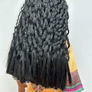 Puede incluir: Una mu&ntilde;eca con pelo largo, negro y rizado. La mu&ntilde;eca lleva un vestido amarillo con un dise&ntilde;o estampado colorido en las mangas y el dobladillo. El pelo est&aacute; peinado de forma voluminosa y texturizada.